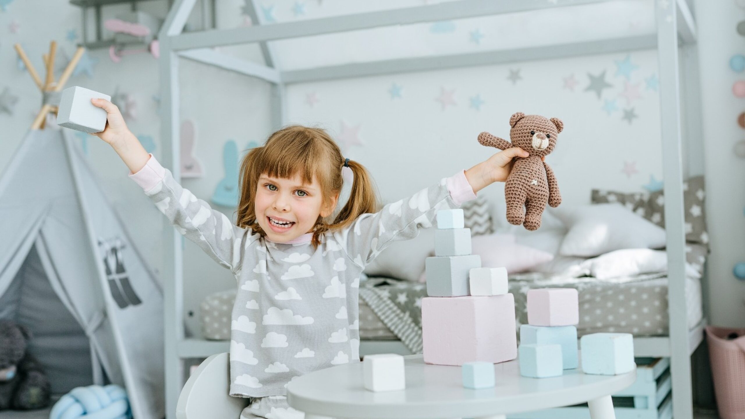 Menina brincando em um quarto infantil decorado com cores suaves, brinquedos e cama estilo cabana ao fundo.
